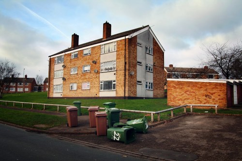 A row of seven upright wheelie bins in various shades of green and brown, positioned on a paved driveway adjacent to a grassy verge, with some bins slightly tilted and open, indicating recent disposal activity. In the background, there is a mid-20th century brick residential building with multiple windows, some open, and a sloped tiled roof, situated on a slight incline with other similar apartment blocks visible further away. The sky above is partly cloudy with patches of blue, and leafless trees are seen to the right, suggesting late autumn or winter. The scene appears to be in an urban or suburban area within the Hammersmith postcode district, consistent with local waste management practices, and the image is relevant to rubbish removal services supplied by Business Waste Removal Hammersmith.