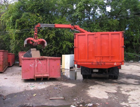 A waste collection scene featuring a red skip loader truck with a large, rectangular, orange waste container attached at the rear. The truck is positioned on an outdoor surface with visible dirt and small debris scattered around. To the left of the truck, a red metal skip with a weathered surface and scratches is seen, partially filled with mixed rubbish materials. A mechanical arm with a claw attachment extends from the truck, actively lifting or emptying waste into the container. In the background, there are tall green trees and foliage, indicating the scene takes place in an open area, possibly near a waste management facility or commercial premises in Hammersmith. The environment appears to be well-lit with natural daylight, highlighting the industrial equipment and the surrounding outdoor ground.