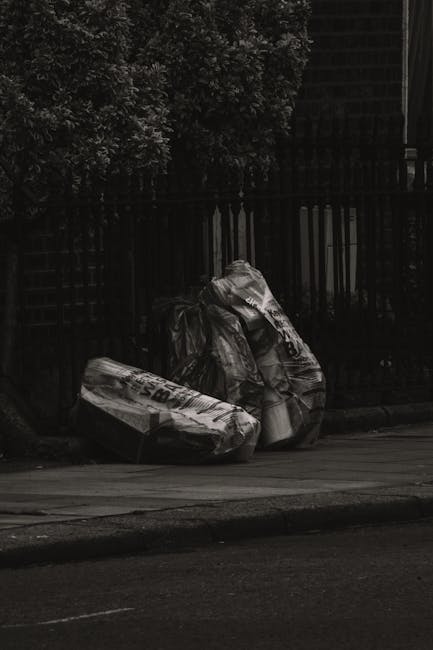 The image depicts a pile of large black plastic rubbish bags placed on the pavement near a black metal fence, with a background of dense foliage and a structure partially visible behind the fence. The