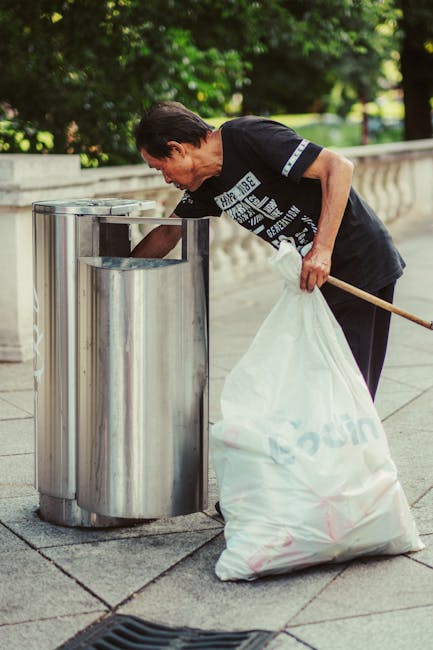 The image depicts a pile of large black plastic rubbish bags placed on the pavement near a black metal fence, with a background of dense foliage and a structure partially visible behind the fence. The