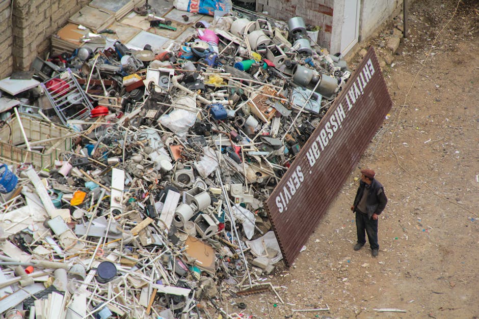 A large pile of assorted rubbish and debris, consisting of broken household appliances, plastic containers, metal frames, and miscellaneous waste materials, is situated on a dirt ground adjacent to a 